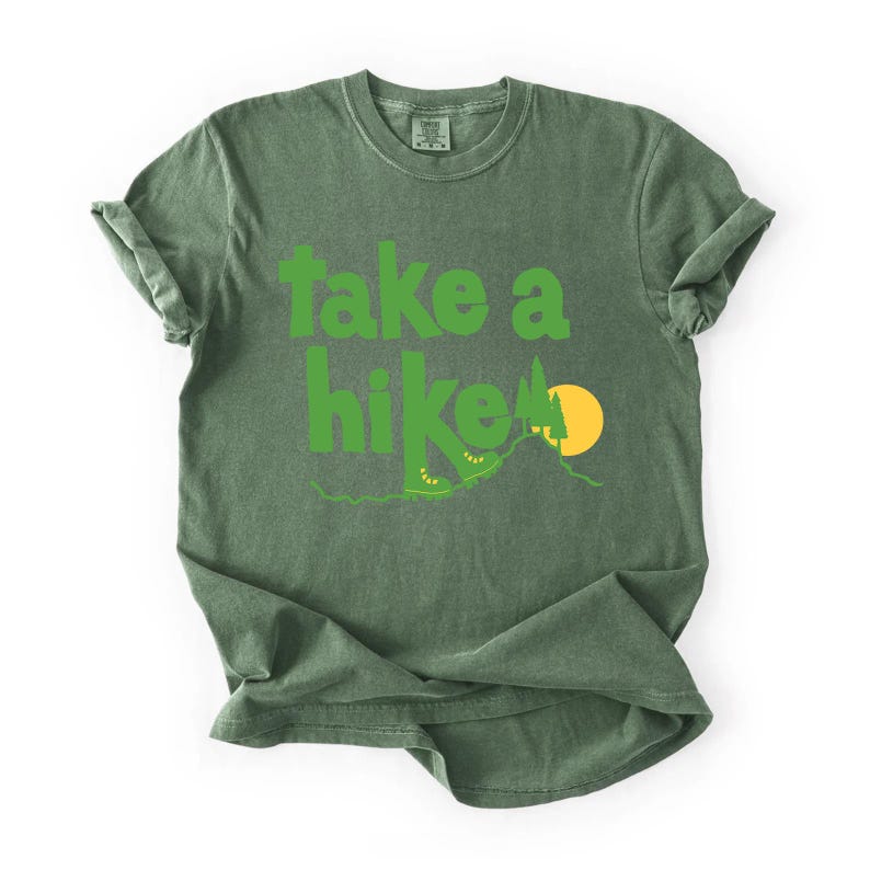 May include: Green t-shirt with the phrase "take a hike" in bright green letters. The design includes hiking boots, a mountain, and a yellow sun. The shirt is a solid green color.