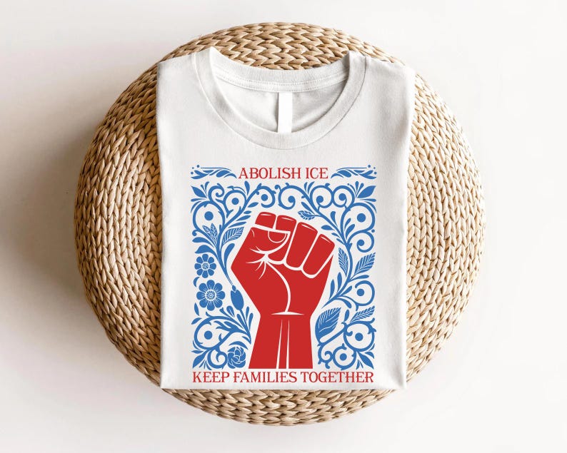 May include: White t-shirt featuring a red fist graphic within a blue floral design. The text "ABOLISH ICE" is above the fist, and "KEEP FAMILIES TOGETHER" is below. The shirt is displayed on a woven, circular surface.