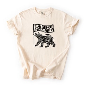 May include: Cream-coloured t-shirt with a black graphic of a bear holding a flag that reads "PROTECT OUR NATIONAL PARKS." The shirt has rolled-up sleeves and a crew neck. The fabric appears soft and comfortable.