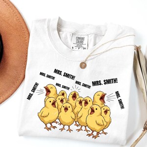 May include: White Comfort Colors t-shirt featuring a graphic of yellow chicks with open beaks, shouting "MRS. SMITH!". The shirt has a crew neck and short sleeves. A gold necklace with a pendant is draped on the shirt.