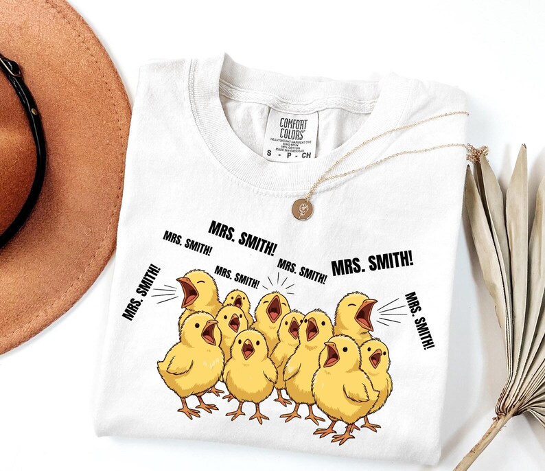 May include: White Comfort Colors t-shirt featuring a graphic of yellow chicks with open beaks, shouting "MRS. SMITH!". The shirt has a crew neck and short sleeves. A gold necklace with a pendant is draped on the shirt.