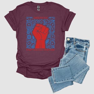 May include: Burgundy t-shirt with a red fist graphic inside a blue floral design. The text on the shirt reads "ABOLISH ICE" and "KEEP FAMILIES TOGETHER." The shirt is paired with blue denim jeans.