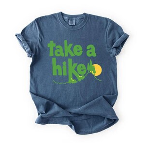 May include: Blue t-shirt with the phrase "take a hike" in green, featuring a hiking boot, mountain, and sun graphic. The shirt is made of a soft material and has rolled-up sleeves. A casual tee for outdoor enthusiasts.