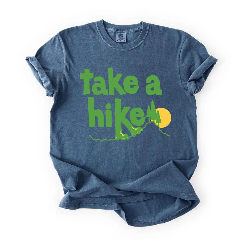 May include: Blue t-shirt with the phrase "take a hike" in green, featuring a hiking boot, mountain, and sun graphic. The shirt is made of a soft material and has rolled-up sleeves. A casual tee for outdoor enthusiasts.