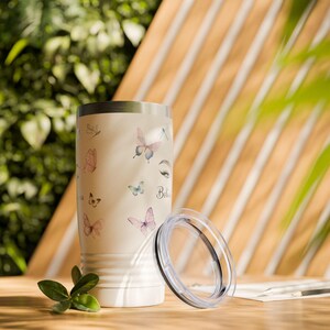Könnte beinhalten: Ein cremefarbener Becher mit Deckel, verziert mit Aquarell-Schmetterlingen in Rosa-, Lila- und Blautönen. Das Wort "Believe" ist auf dem Becher geschrieben. Der Becher steht auf einer Holzoberfläche.