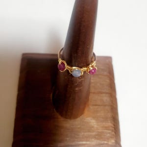 May include: A gold-colored wire-wrapped ring with three gemstones. The ring features two red gemstones and one light blue gemstone. The ring is displayed on a wooden ring holder.