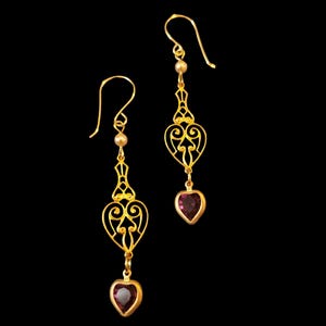 May include: A pair of gold-toned drop earrings featuring a filigree heart-shaped metal component suspended from a hook wire. Each earring includes a small round bead accent and a faceted, heart-shaped dark purple gemstone dangling at the bottom.