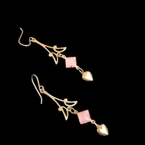 Può includere: Un paio di orecchini pendenti color oro con una sezione superiore decorativa a forma di ramo, una pietra quadrata rosa chiaro e un piccolo ciondolo a forma di cuore color oro alla base. Gli orecchini sono appesi a ganci su sfondo nero.