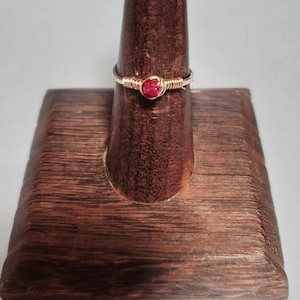 May include: A gold-toned wire-wrapped ring with a small, round, red gemstone. The ring is displayed on a wooden ring holder with a square base and a tapered top. The gemstone is the focal point, set in the center of the ring.