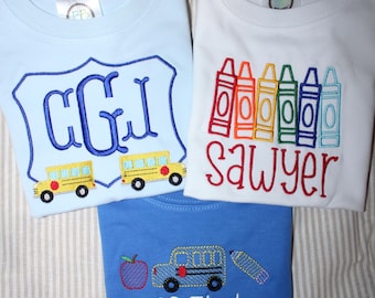 May include: Three t-shirts with school-themed embroidery. One light blue shirt features a blue monogram and two yellow school buses. One white shirt displays colorful crayon graphics and the name Sawyer. One blue shirt shows an apple, bus, and pencil.