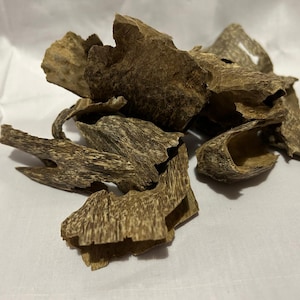 以下が含まれることがあります： 薄茶色の、層状に見える木片の山。木片は自然で有機的な外観をしており、形や大きさが様々です。背景は白い表面です。