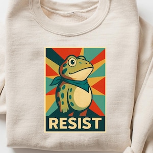 May include: Cream-colored sweatshirt featuring a cartoon frog with a blue bandana, set against a retro-style background with the word "RESIST". The frog is green with yellow spots. The background has red, yellow, and teal stripes.
