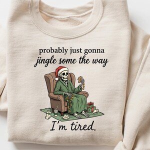 K&ouml;nnte beinhalten: Beiges Sweatshirt mit dem Text "probably just gonna jingle some the way, I'm tired." Die Grafik zeigt ein Skelett mit Weihnachtsm&uuml;tze, das in einem Stuhl sitzt, umgeben von Geschenken. Ein humorvolles Design zum Thema Weihnachten.