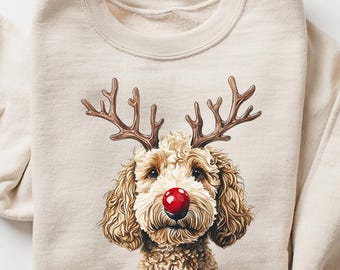 May include: A beige sweatshirt featuring a dog with reindeer antlers and a red nose. The dog's face is the focal point, with detailed fur and a festive holiday design. The sweatshirt is folded, with the sleeves casually draped.