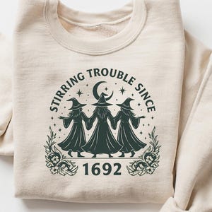 May include: Beige sweatshirt with a circular graphic featuring three witch figures in dark green robes and hats, holding hands under a crescent moon and stars. The text reads "Stirring Trouble Since 1692."