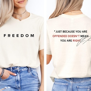 May include: Cream-coloured t-shirt with the word "FREEDOM" printed in black on the front. The back of the shirt features the quote, "JUST BECAUSE YOU ARE OFFENDED DOESN'T MEAN YOU ARE RIGHT" in black and red text, with a signature.