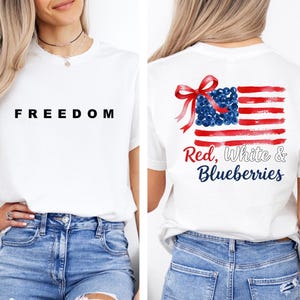 May include: White t-shirt with the word "FREEDOM" in black block letters on the front. The back of the shirt features a watercolor-style American flag design with the text "Red, White & Blueberries".