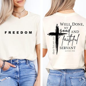 May include: A cream-coloured t-shirt with the word "FREEDOM" printed in black on the front. The back of the shirt features a black cross design with the text "Well Done, My Good and Faithful Servant" and "Matthew 25:21".