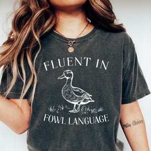 May include: A dark gray t-shirt with a white graphic of a duck and the text "Fluent in Fowl Language".
