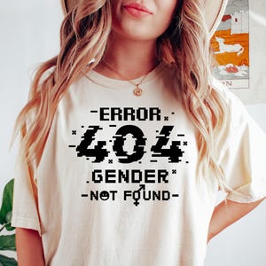 May include: A white t-shirt with black text that reads "ERROR 404 .GENDER. -NOT FOUND-." The text is in a pixelated font, giving it a glitch effect.