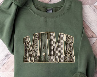 May include: An olive green sweatshirt with the word "MAMA" in a collegiate-style font. The letters have a patterned design with a mix of leopard print and checkered patterns. The sweatshirt is folded on a light wood-grain surface.