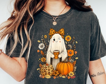 May include: Dark gray t-shirt featuring a cute ghost character holding a coffee cup, surrounded by pumpkins and fall foliage. The ghost wears a bow. A fun Halloween-themed graphic tee.