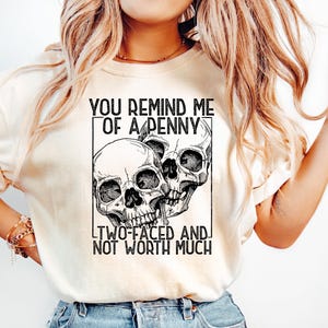 May include: Cream-colored t-shirt featuring a black and white graphic of three skulls within a rectangular frame. The text above reads "YOU REMIND ME OF A PENNY," and below, "TWO-FACED AND NOT WORTH MUCH."