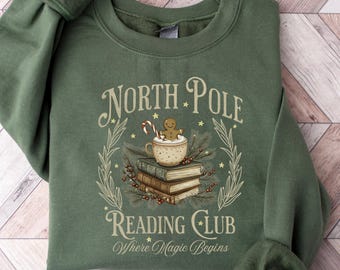 May include: Green sweatshirt with the text "North Pole Reading Club" and an illustration of a gingerbread man, books, and a cup of cocoa. The phrase "Where Magic Begins" is also included. The sweatshirt is laid flat on a wooden surface.