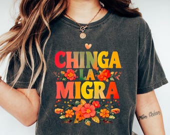 May include: Dark gray t-shirt with the text "CHINGA LA MIGRA" in colorful letters, decorated with floral elements. The shirt is paired with blue jeans and white sneakers.