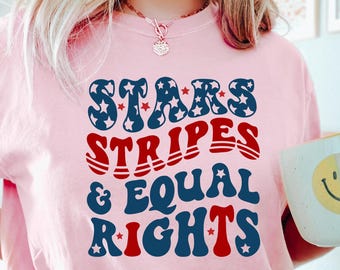 May include: A light pink t-shirt with the words "STARS STRIPES & EQUAL RIGHTS" in a retro font. The text is in red and blue, with star accents. A white mug with a yellow smiley face is held in the hand.
