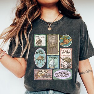 May include: Dark gray t-shirt featuring a collection of whimsical postage stamp designs. The stamps display various space-themed illustrations and phrases like "Why Stupid?" and "I am scary space monster."