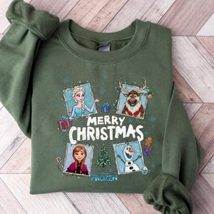 Puede incluir: Una sudadera verde oliva con personajes de la pel&iacute;cula Frozen. El dise&ntilde;o incluye a Elsa, Anna, Olaf y Sven, con las palabras "Merry Christmas" y la palabra "Frozen" debajo. La sudadera est&aacute; sobre una superficie de madera.