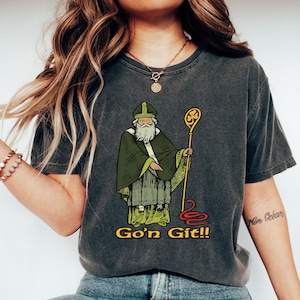 May include: Dark gray t-shirt featuring a cartoon illustration of a saintly figure in green robes and a staff. The text "Go'n Git!!" is in yellow. The shirt is a casual wear item.