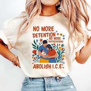 May include: Cream-coloured t-shirt with the text "NO MORE DETENTION, NO MORE DEPORTATIONS, ABOLISH I.C.E." in a colourful floral design. The graphic includes a family illustration. The person in the image is wearing light blue jeans.