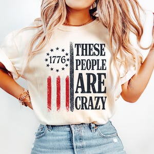 May include: Cream-colored t-shirt with a distressed American flag design. The flag features red and black stripes, a star-studded section with "1776," and the text "THESE PEOPLE ARE CRAZY" in bold black letters. The shirt is paired with light blue jeans.