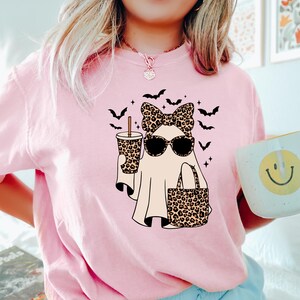 May include: Pink t-shirt featuring a cartoon ghost wearing sunglasses, a bow, and carrying a leopard print bag and drink. Black bats are flying above the ghost. A white mug with a yellow smiley face is held in the hand.