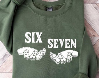 May include: An olive green sweatshirt with the words "SIX SEVEN" in white block letters. Below the text are two white illustrations of hands. The sweatshirt is folded on a light wood grain surface.