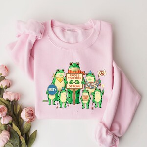 Puede incluir: Sudadera rosa claro con un dise&ntilde;o de ranas verdes de dibujos animados. Las ranas sostienen carteles con las frases "Unity", "Frogs Together Strong" y "No Hope Without". Las ranas llevan accesorios como pa&ntilde;uelos y sombreros.