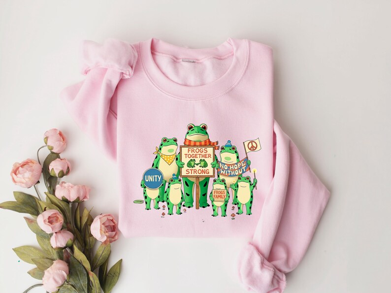 Puede incluir: Sudadera rosa claro con un dise&ntilde;o de ranas verdes de dibujos animados. Las ranas sostienen carteles con las frases "Unity", "Frogs Together Strong" y "No Hope Without". Las ranas llevan accesorios como pa&ntilde;uelos y sombreros.