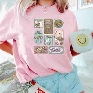 May include: A light pink t-shirt featuring a collection of whimsical postage stamp designs. The stamps include phrases like "Why Stupid?" and "I am scary space monster." The shirt is paired with light blue shorts and a mug with a smiley face.