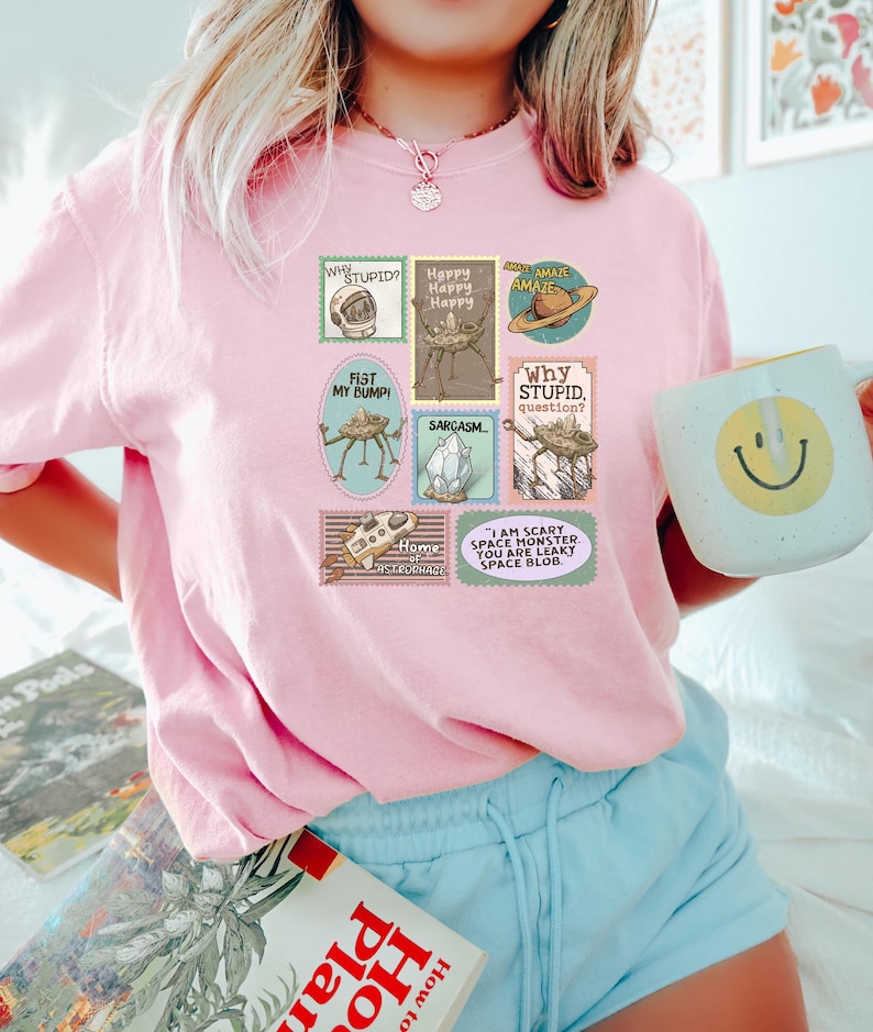 May include: A light pink t-shirt featuring a collection of whimsical postage stamp designs. The stamps include phrases like "Why Stupid?" and "I am scary space monster." The shirt is paired with light blue shorts and a mug with a smiley face.