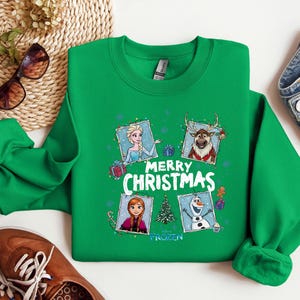 Puede incluir: Una sudadera verde de cuello redondo con un dise&ntilde;o festivo con personajes de la pel&iacute;cula Frozen. El dise&ntilde;o incluye a Elsa, Anna, Olaf y Sven, con las palabras "Merry Christmas" en blanco. La sudadera tiene mangas largas y pu&ntilde;os acanalados.
