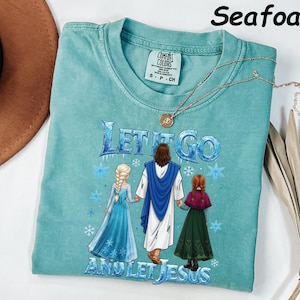 May include: Seafoam green t-shirt with the text "Let It Go and Let Jesus" and a graphic of Jesus with two characters from the movie Frozen. The shirt has a crew neck and short sleeves. A gold necklace with a pendant is draped on the shirt.