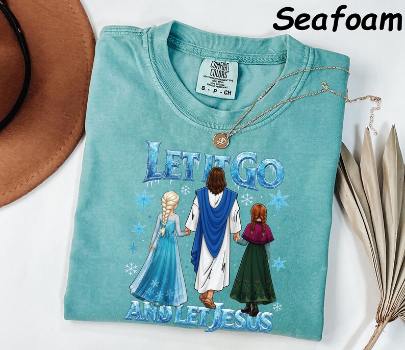 May include: Seafoam green t-shirt with the text "Let It Go and Let Jesus" and a graphic of Jesus with two characters from the movie Frozen. The shirt has a crew neck and short sleeves. A gold necklace with a pendant is draped on the shirt.