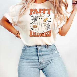 May include: Cream-colored t-shirt with the words "PAPPY HALLOWEEN" in orange block letters. A cartoon ghost wearing a nurse's hat and holding a syringe and a pumpkin is in the center. Black bats and spiderwebs surround the ghost.