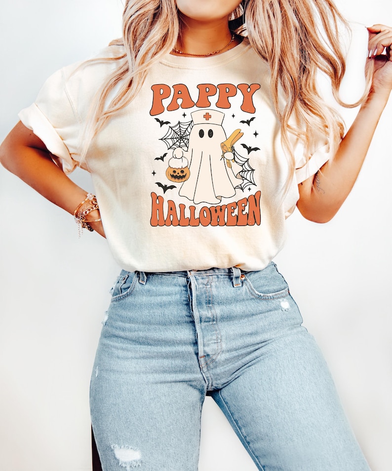 May include: Cream-colored t-shirt with the words "PAPPY HALLOWEEN" in orange block letters. A cartoon ghost wearing a nurse's hat and holding a syringe and a pumpkin is in the center. Black bats and spiderwebs surround the ghost.