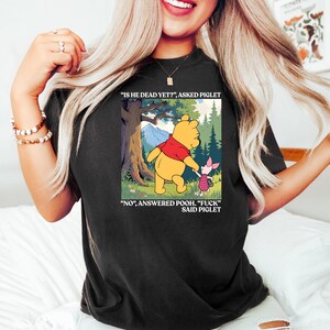 Puede incluir: Camiseta negra con una imagen de dibujos animados de Winnie the Pooh y Piglet. La imagen incluye el texto "IS HE DEAD YET?" y "NO", ANSWERED POOH. "FUCK" SAID PIGLET". Es una prenda informal.