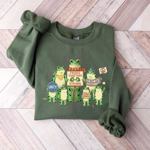 Puede incluir: Sudadera verde con un gr&aacute;fico de ranas de dibujos animados sosteniendo carteles. Los carteles dicen "Frogs Together Strong" y "No Hope Without Frogs". Las ranas est&aacute;n en diferentes poses, algunas con pa&ntilde;uelos y s&iacute;mbolos de paz.