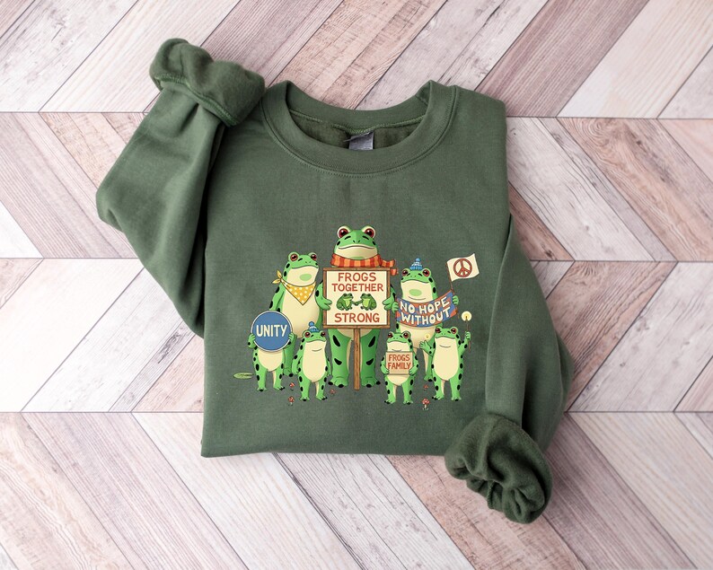 Puede incluir: Sudadera verde con un gr&aacute;fico de ranas de dibujos animados sosteniendo carteles. Los carteles dicen "Frogs Together Strong" y "No Hope Without Frogs". Las ranas est&aacute;n en diferentes poses, algunas con pa&ntilde;uelos y s&iacute;mbolos de paz.