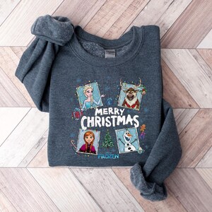 Puede incluir: Una sudadera gris oscuro con personajes de la pel&iacute;cula Frozen. La sudadera muestra las palabras "Merry Christmas" e im&aacute;genes de Elsa, Anna, Olaf y Sven. La sudadera est&aacute; hecha de un material suave, ideal para la temporada navide&ntilde;a.